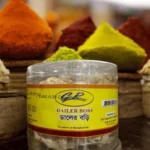 A clear jar of G.R. brand Dailer Bori, traditional sun-dried lentil dumplings, with colorful spice mounds in the background.