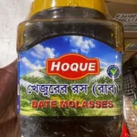 hoque-date-molasses-khejur-gur A person holding a large jar of Hoque Date Molasses against a store background, showing its size and clear labeling.