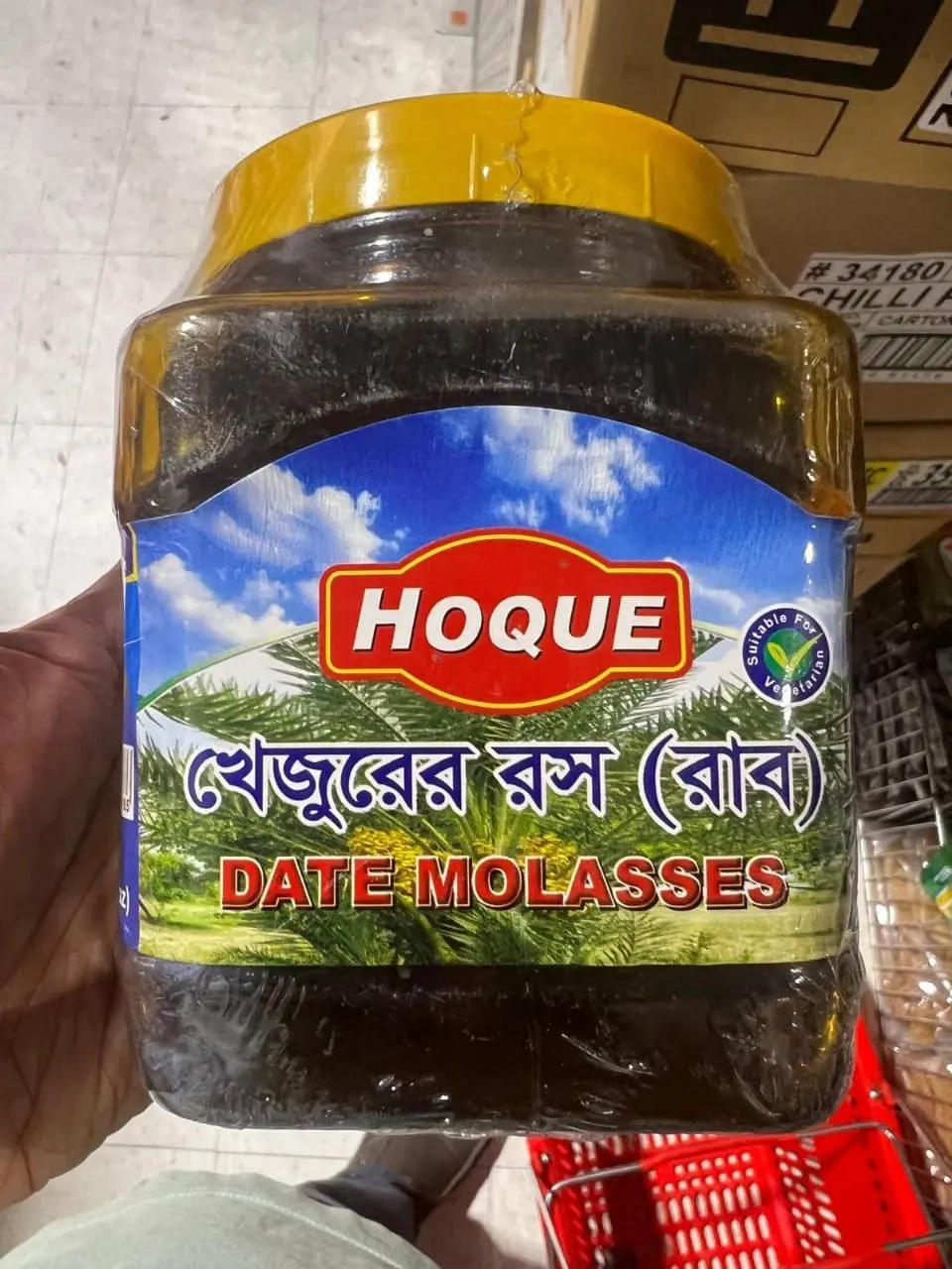 hoque-date-molasses-khejur-gur A person holding a large jar of Hoque Date Molasses against a store background, showing its size and clear labeling.